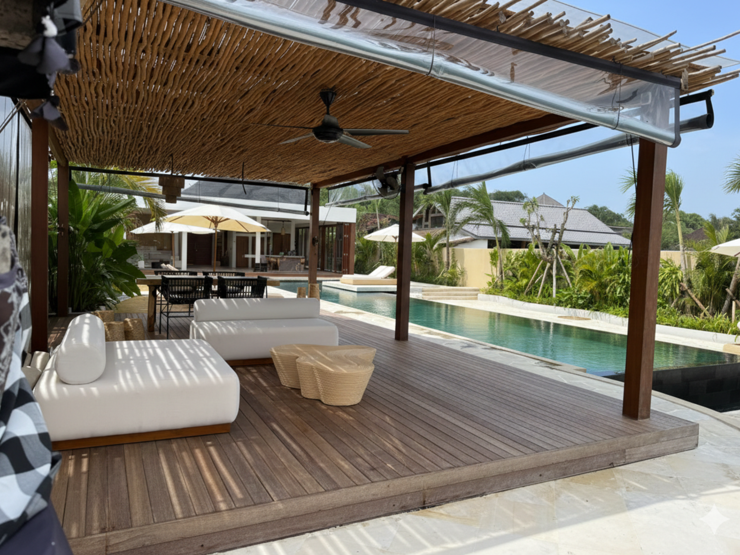 Overdekt houten terras met witte daybeds onder een bamboe pergola naast het zwembad; achteraan staan een eettafel en parasols