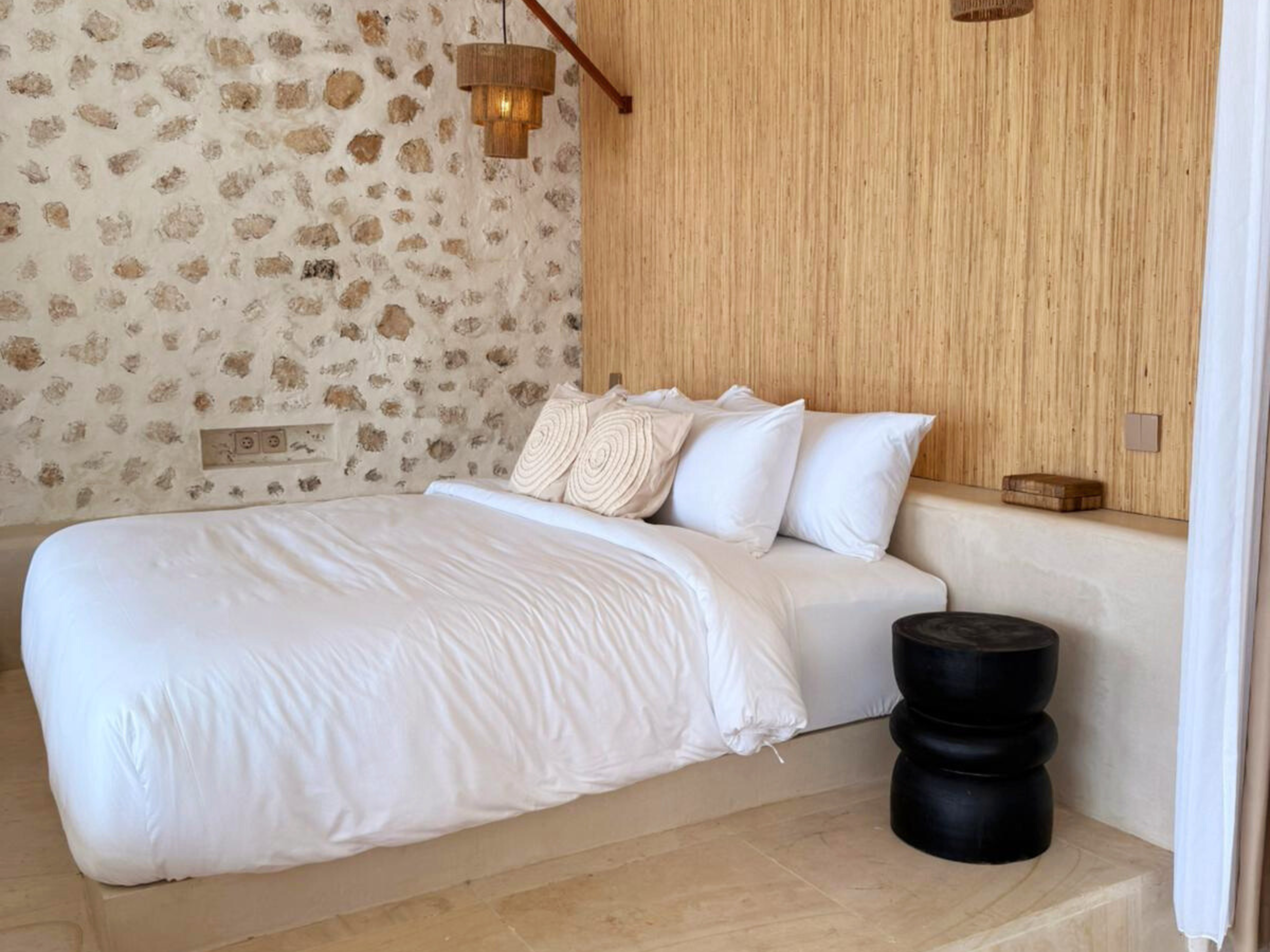 Slaapkamer met wit beddengoed, stenen muur, bamboe wandbekleding en hanglamp