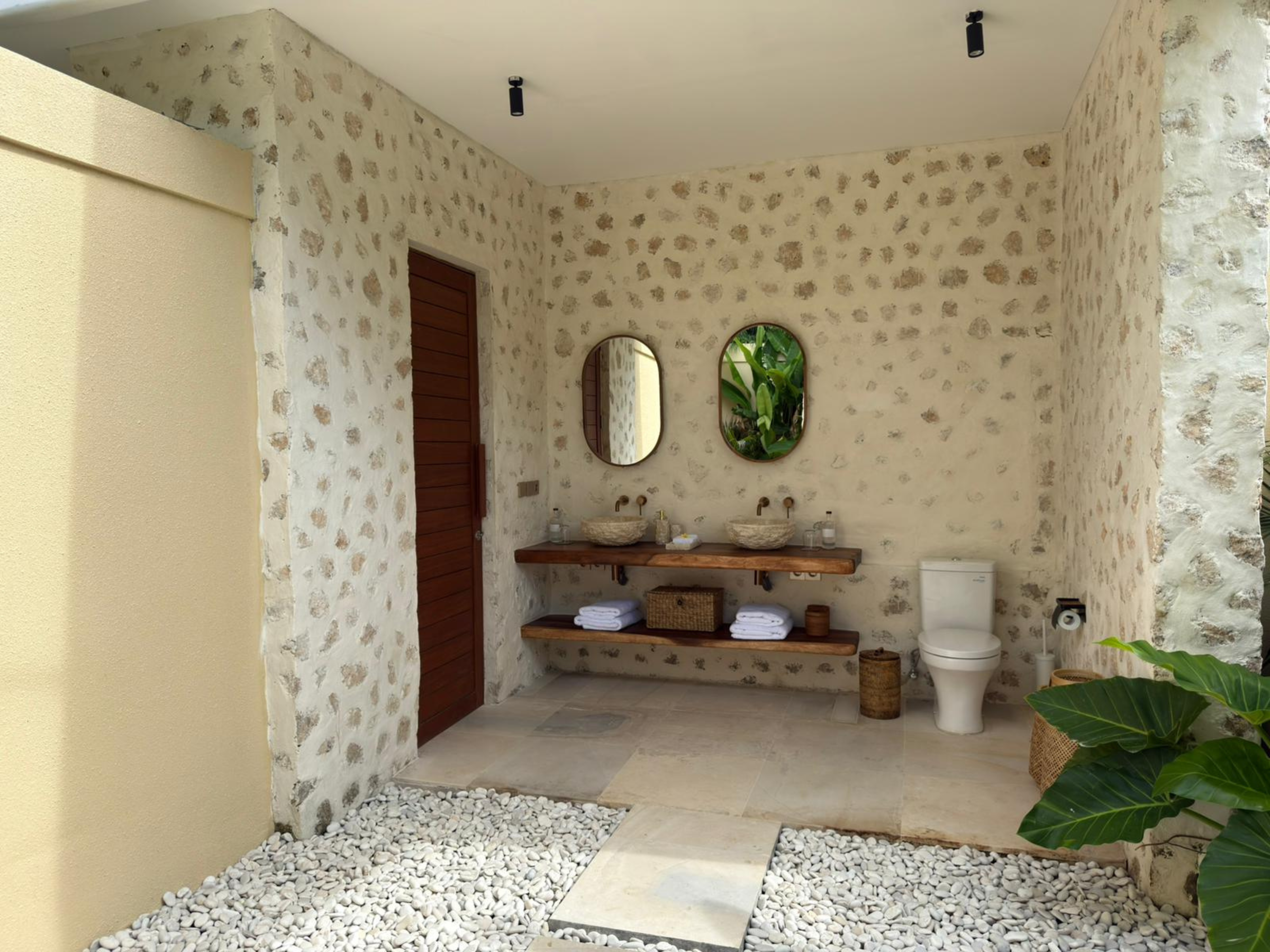 Open badkamer met natuurstenen muren, houten deur, twee ronde spiegels en houten wasmeubel; rechts een toilet en links een plant