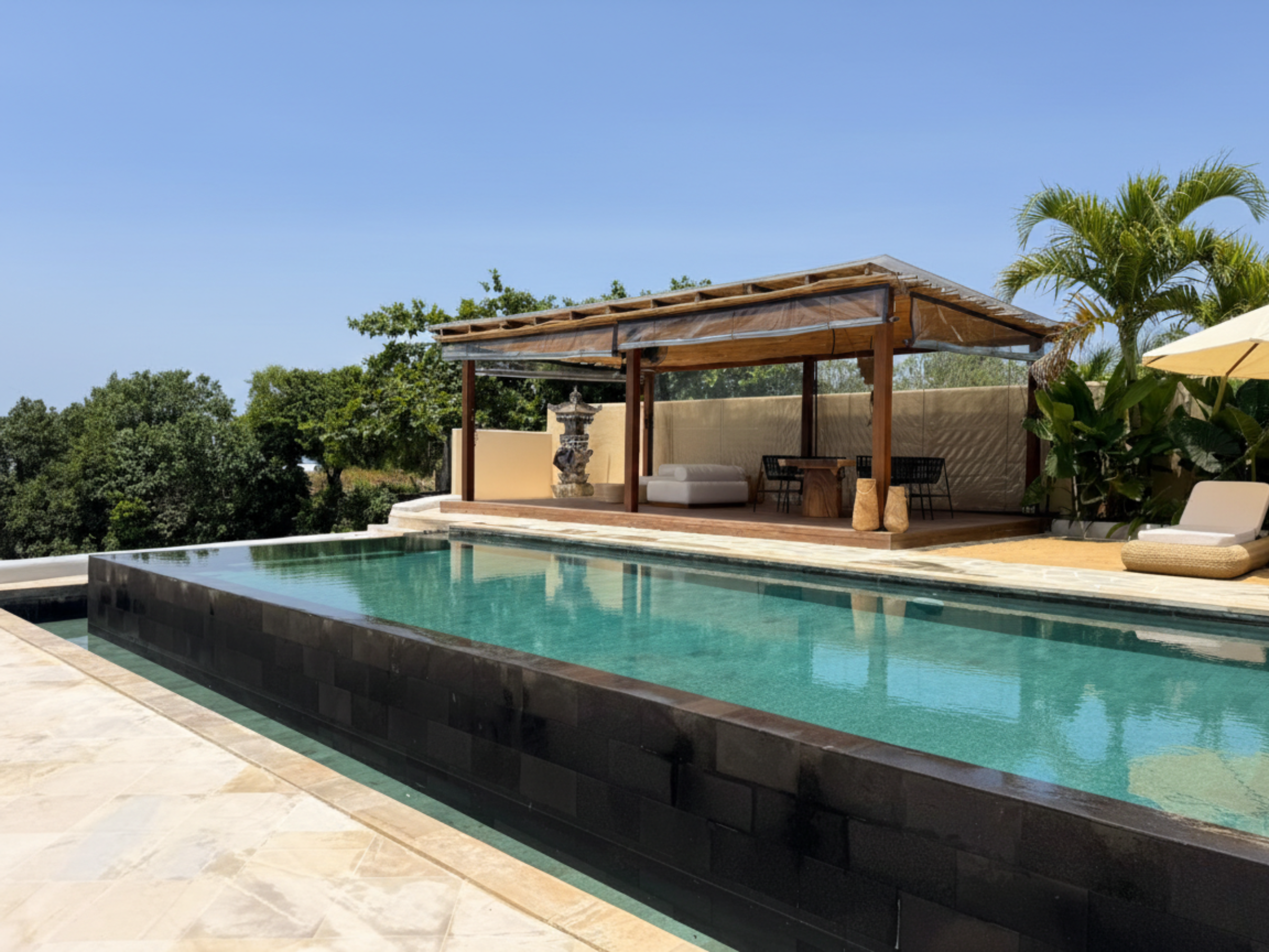 Infinity‑zwembad met donkere stenen rand en een open loungepaviljoen; rechts een ligbed en parasol
