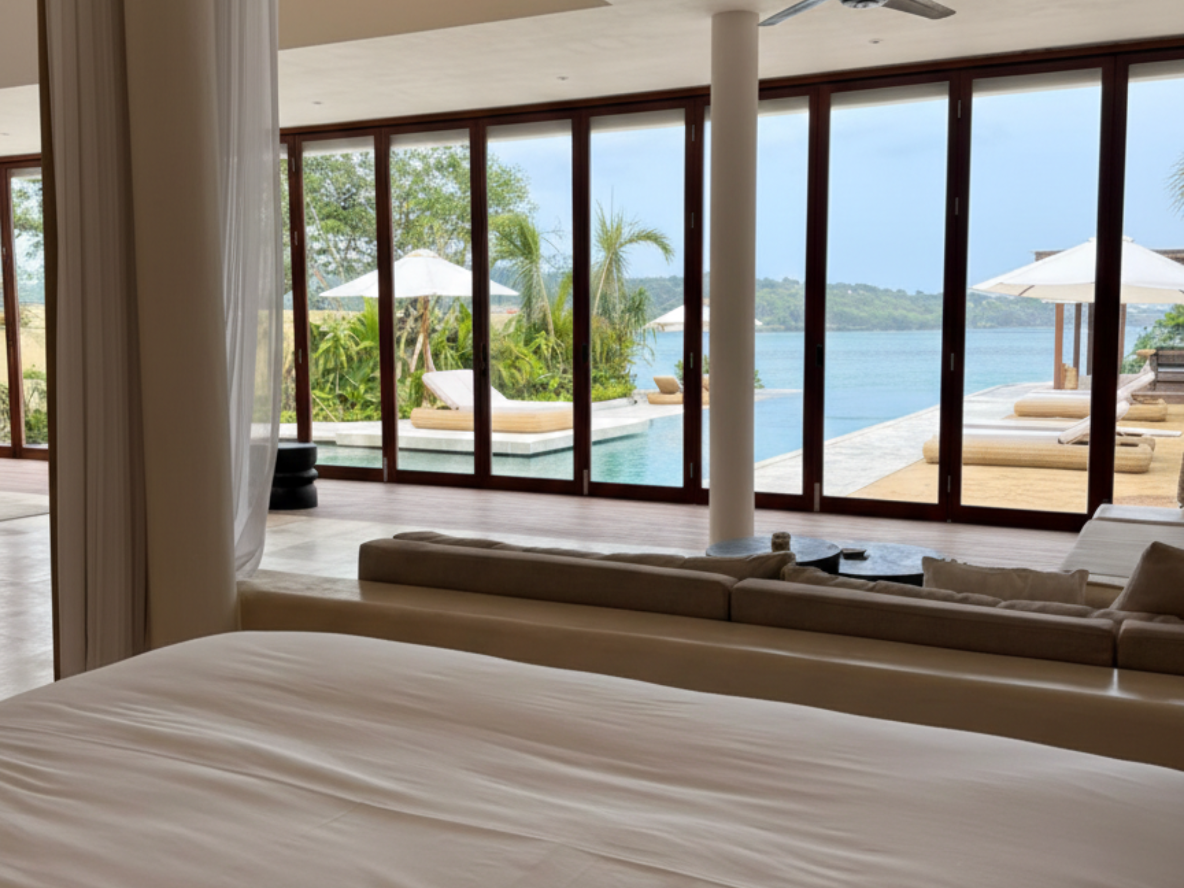 Binnenzicht vanuit een slaapkamer met hemelbed en grote sofa; glazen schuifdeuren openen naar het zwembad en de zee