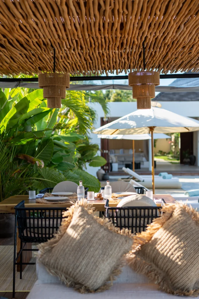 Overdekt houten terras met witte daybeds onder een bamboe pergola naast het zwembad; achteraan staan een eettafel en parasols