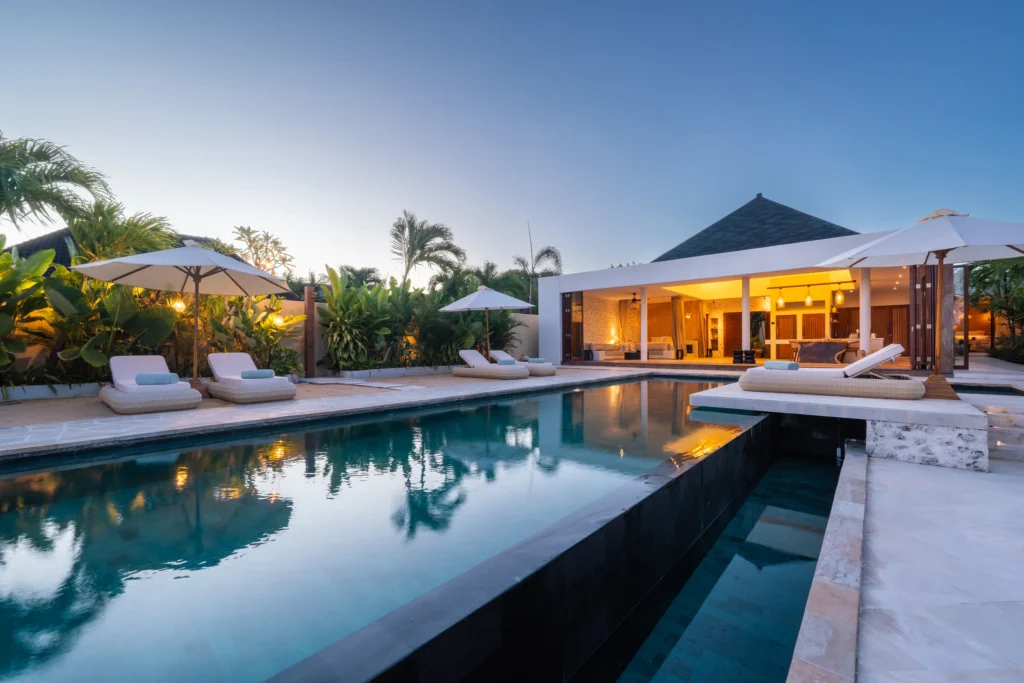 Infinity‑zwembad met donkere stenen rand en een open loungepaviljoen; rechts een ligbed en parasol