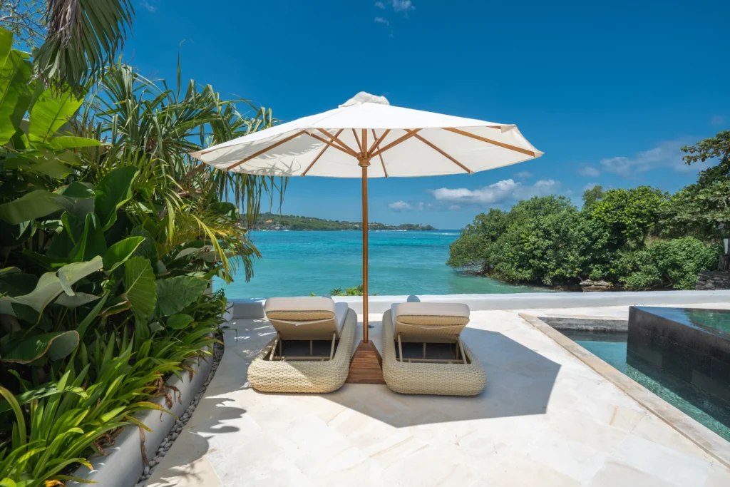 Zwembad gezien vanaf de andere zijde; langs beide kanten loungebedden met parasols en op de achtergrond de villa