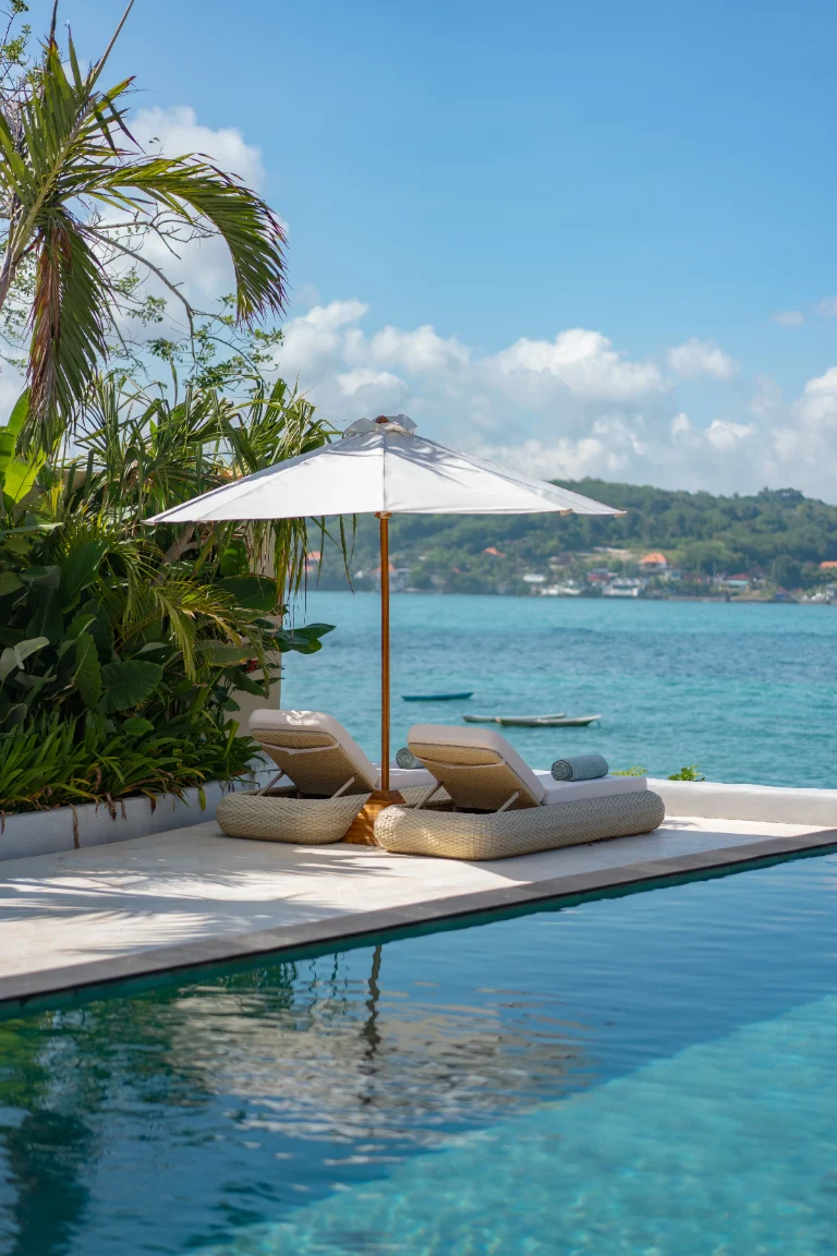 Daybeds under a parasol at the pool's edge, boat drifting on the turquoise bay