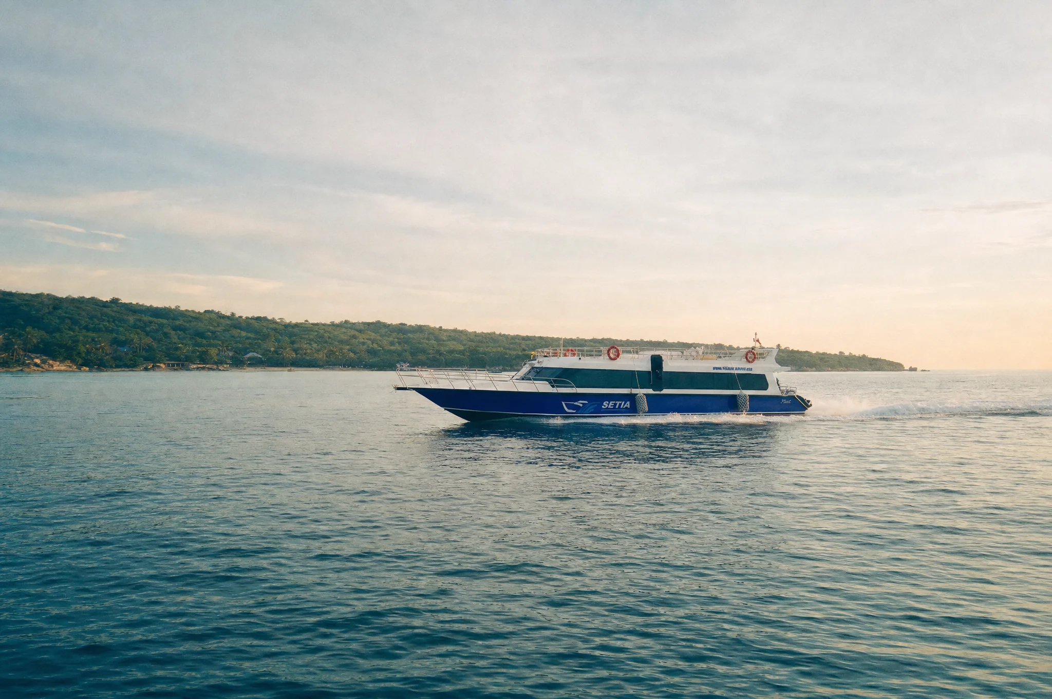 Blue and white Setia fast boat crossing to Nusa Lembongan at golden hour, forested coastline behind